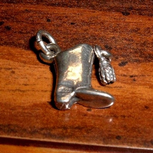 May include: A silver-toned charm in the shape of a cowboy boot with a loop for attaching to a bracelet or necklace. The boot has detailed textures and a small, attached spur.