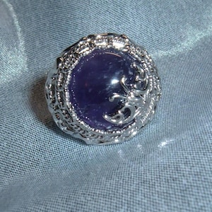 May include: A silver-toned ring with a large, round, deep purple stone at the center. The ring's setting features an ornate, textured design around the stone. The ring is set against a light blue fabric background.
