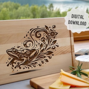 Könnte beinhalten: Ein helles Holzschneidebrett mit einem dekorativen Froschdesign. Der Frosch ist von wirbelnden floralen Elementen umgeben. Die Worte "DIGITALER DOWNLOAD" stehen in einer weißen Blase. Käsescheiben liegen auf einem kleineren Holzbrett.