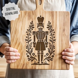 Pode incluir: Uma tábua de cortar de madeira com um design gravado a laser de um quebra-nozes de pé em uma coroa de flores estilizadas. O quebra-nozes está usando um chapéu e um uniforme tradicionais.