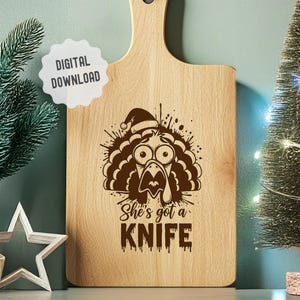 May include: Wooden cutting board with a handle, featuring a turkey design wearing a Santa hat. The text "She's got a KNIFE" is etched below the turkey. A "DIGITAL DOWNLOAD" sticker is in the upper left corner. The board is light brown.