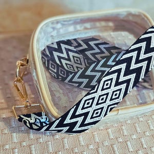 May include: A clear, square crossbody bag with a black and white geometric patterned strap. The strap features a repeating zigzag and diamond design. The bag has gold-colored hardware and a beige trim.