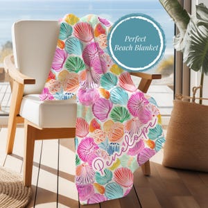 May include: A beach blanket with a seashell pattern in shades of pink, orange, blue, and green is draped over a white chair. The blanket has the text "Penelope" in cursive and a blue circle with the text "Perfect Beach Blanket".