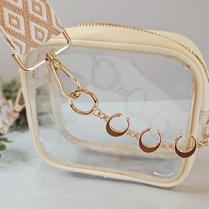 May include: A clear, cream-colored crossbody bag with a gold zipper and hardware. The bag has a patterned strap in cream and tan. A gold chain with crescent moon charms hangs from the bag.