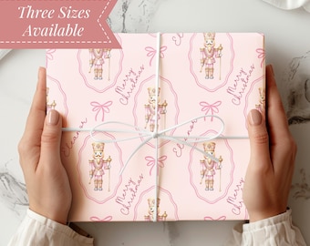 May include: Pink wrapping paper with a repeating pattern of nutcracker soldiers, bows, and the words "Merry Christmas" and "Eleanor." The paper is tied with white string and has a banner that says "Three Sizes Available."