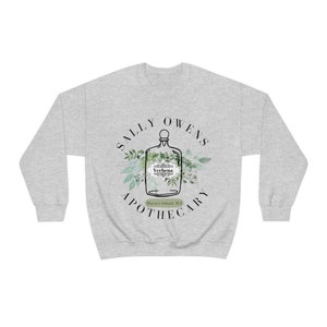 Może przedstawiać: Szara bluza z białym nadrukiem przedstawiającym butelkę z etykietą z napisem "Verbena" oraz tekstem "Sally Owens Apothecary, Morris Island, MA".