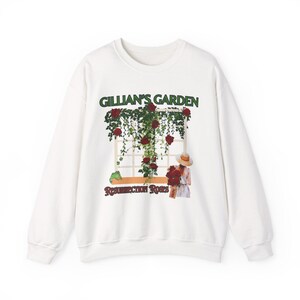 Pode incluir: Camisola branca com um gráfico com uma janela com rosas trepadeiras e o texto "Gillian's Garden" em verde acima de "Resurrection Roses" em castanho. Uma pessoa com um chapéu segura rosas.