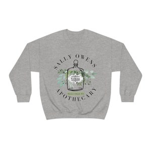 Może przedstawiać: Szara bluza z czarno-białym wzorem przedstawiającym butelkę z etykietą z napisem "Verbena" i tekstem "Sally Owens Apothecary" wok&oacute;ł butelki. Tekst "Maris's Island, MA" znajduje się pod butelką.