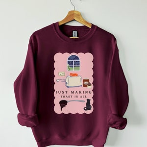 May include: A maroon sweatshirt with a pink graphic of a toaster, bread, and a cat. The text "JUST MAKING TOAST IS ALL" is printed on the graphic.