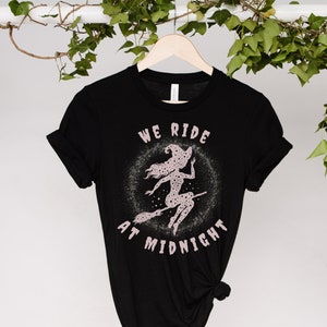 May include: Black t-shirt with a pink graphic of a witch flying on a broomstick with stars and the text "We Ride At Midnight".
