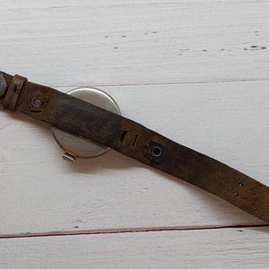 May include: A vintage wristwatch featuring a distressed brown leather strap and a silver watch face. The strap shows signs of wear and age, with a metal buckle. The watch is set against a light-coloured wooden surface.