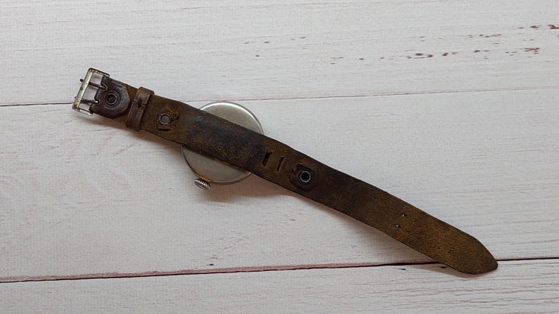 May include: A vintage wristwatch featuring a distressed brown leather strap and a silver watch face. The strap shows signs of wear and age, with a metal buckle. The watch is set against a light-coloured wooden surface.