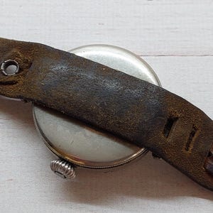 May include: A close-up of a vintage wristwatch featuring a silver face and a distressed brown leather strap. The strap shows wear and tear, with visible stitching and metal fixings. The watch's winder is visible.
