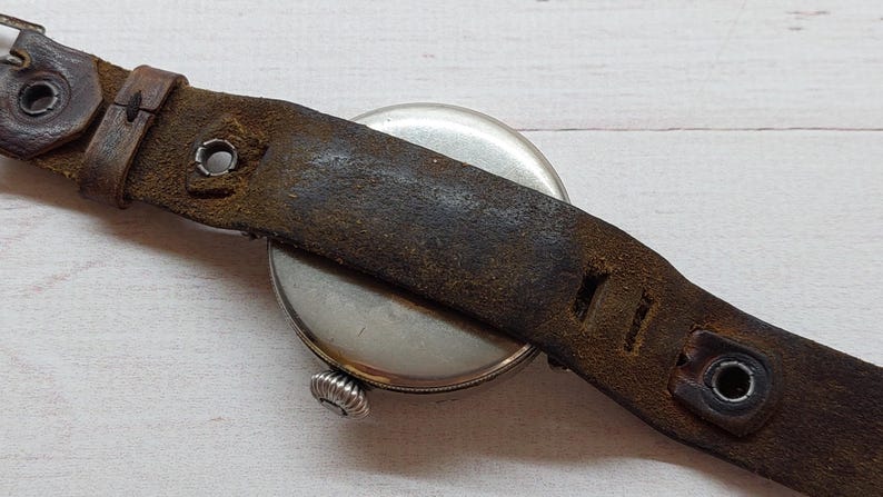 May include: A close-up of a vintage wristwatch featuring a silver face and a distressed brown leather strap. The strap shows wear and tear, with visible stitching and metal fixings. The watch's winder is visible.