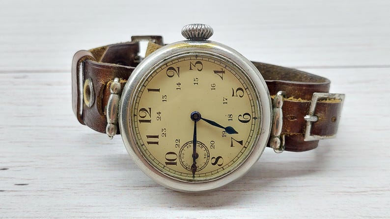 May include: A vintage silver wristwatch with a cream face and black numerals. The watch features a small seconds dial and is attached to a worn brown leather strap. The watch face displays the numbers 1 to 24.