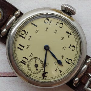 May include: A vintage wristwatch with a cream face, black numerals, and blue hands. The watch has a silver case and a brown leather strap. A small seconds dial is at the bottom of the face.