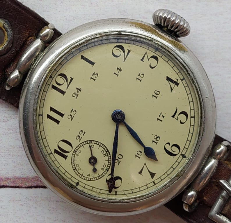 May include: A vintage wristwatch with a cream face, black numerals, and blue hands. The watch has a silver case and a brown leather strap. A small seconds dial is at the bottom of the face.