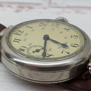May include: A close-up of a vintage wristwatch with a cream-coloured face and black numerals. The watch has blue hands and a small subdial. It is attached to a brown leather strap. The watch case is silver-coloured.