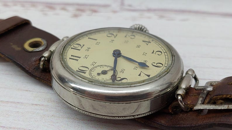 May include: A close-up of a vintage wristwatch with a cream-coloured face and black numerals. The watch has blue hands and a small subdial. It is attached to a brown leather strap. The watch case is silver-coloured.