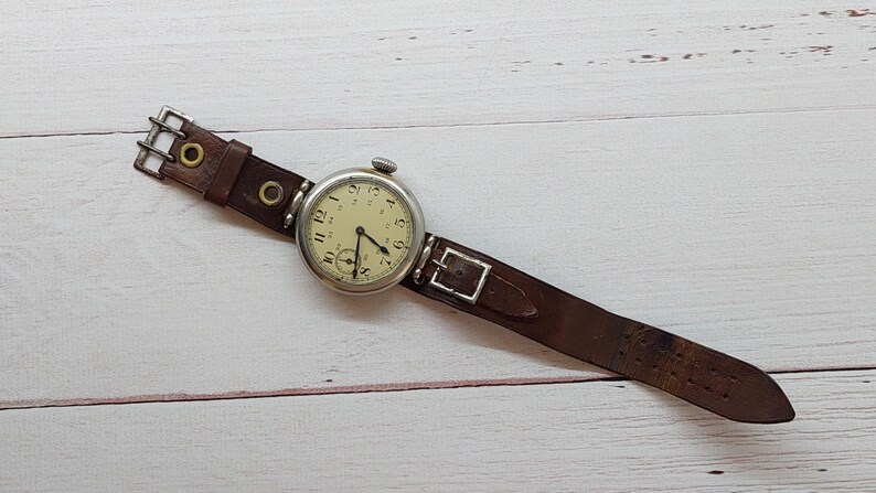 May include: A vintage wristwatch with a cream-coloured face, black numerals, and black hands. The watch has a silver-coloured case and a brown leather strap with a buckle. The watch is lying on a light-coloured wooden surface.