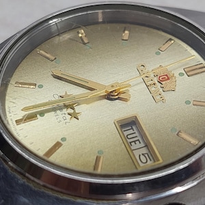 Può includere: Primo piano di un orologio Orient vintage con cinturino in metallo argentato. Il quadrante dell'orologio è dorato con lancette e indicatori dorati. Il giorno è visualizzato come "MAR 5". L'orologio ha un design classico e retrò.