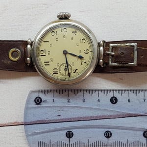 May include: A vintage wristwatch featuring a cream dial with black numerals and a subdial. The watch has a silver case and a brown leather strap with a buckle. A ruler is placed below the watch.