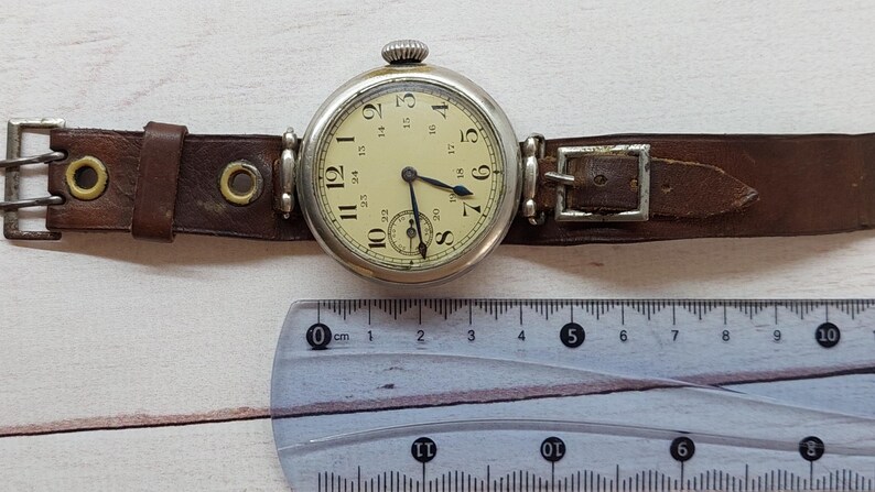 May include: A vintage wristwatch featuring a cream dial with black numerals and a subdial. The watch has a silver case and a brown leather strap with a buckle. A ruler is placed below the watch.