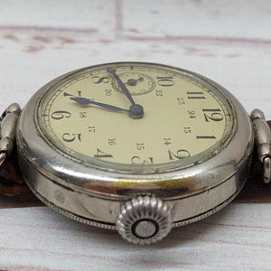 May include: A close-up shot of a vintage wristwatch. The watch has a cream-coloured face with black numerals and a silver-coloured case. It features a brown leather strap. The time on the watch is approximately 10:10.