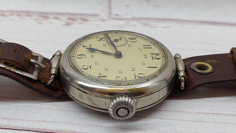 May include: A close-up shot of a vintage wristwatch. The watch has a cream-coloured face with black numerals and a silver-coloured case. It features a brown leather strap. The time on the watch is approximately 10:10.