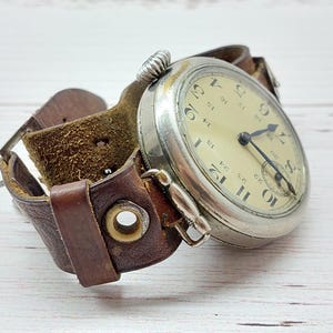 May include: A vintage pocket watch with a silver case and a cream-coloured face, featuring black numerals and hands. The watch is attached to a wide, brown leather cuff-style strap. The strap has a buckle and metal eyelets.