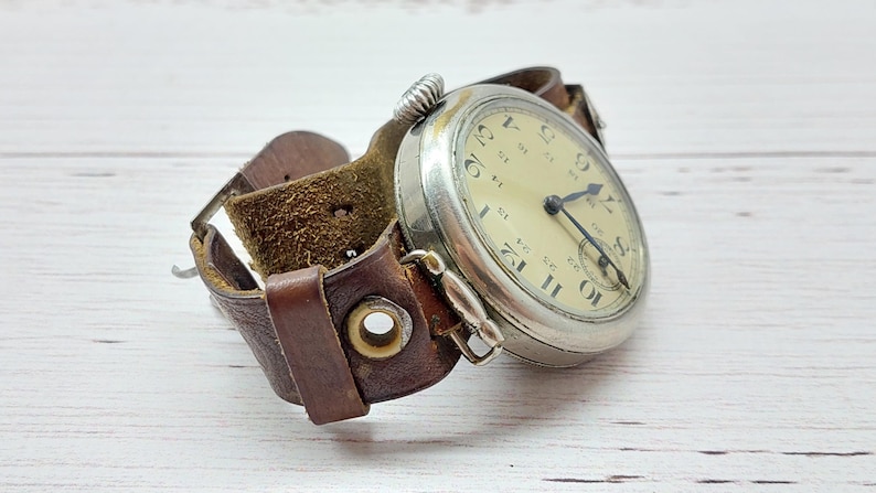 May include: A vintage pocket watch with a silver case and a cream-coloured face, featuring black numerals and hands. The watch is attached to a wide, brown leather cuff-style strap. The strap has a buckle and metal eyelets.