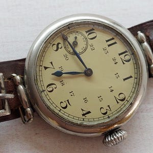May include: A vintage wristwatch with a silver case and a cream face, displaying black numerals and blue hands. It features a small subdial and is secured by a brown leather strap with a metal buckle. The watch has a classic design.