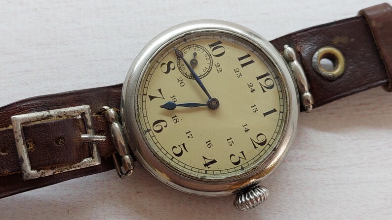 May include: A vintage wristwatch with a silver case and a cream face, displaying black numerals and blue hands. It features a small subdial and is secured by a brown leather strap with a metal buckle. The watch has a classic design.