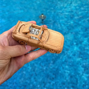 Puede incluir: Un coche de juguete de madera con techo descapotable, sostenido contra un fondo de agua azul. El coche está hecho de madera de color claro y tiene detalles, incluyendo ruedas y un interior abierto. El coche mide aproximadamente 15 cm de largo.