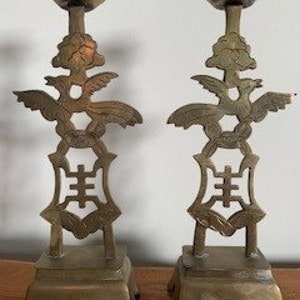Könnte beinhalten: Ein Paar verzierte Kerzenleuchter aus Messing mit einem Lotusblumen-Design auf der Oberseite und einem Vogel- und Blumenmotiv in der Mitte. Die Kerzenleuchter haben einen quadratischen Sockel mit aufwendigen Details.