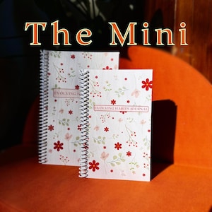 May include: Two spiral bound journals with a white background and a floral pattern in red, pink, and green. The text "Evolving Habits Journal" is printed on the front of the journal.