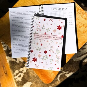 May include: A white and black spiral bound journal with a floral pattern and the text "Evolving Habits Journal". The journal is open to a page with the title "Rate My Day" and a grid for rating the day on a scale of 1 to 5 stars.