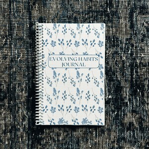 May include: A white spiral bound journal with a blue floral pattern and the text "Evolving Habits Journal" on the front cover.