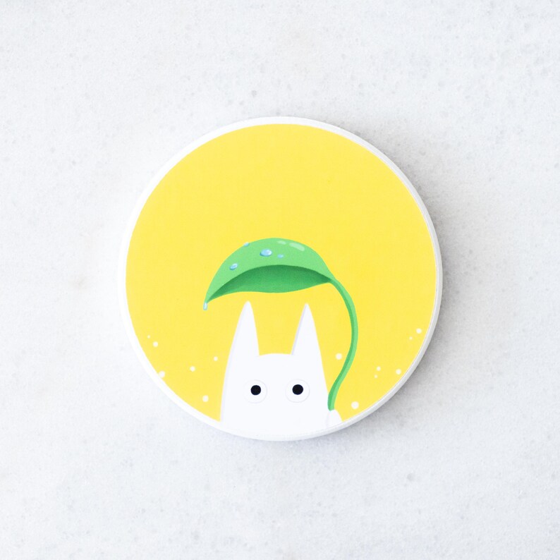 Puede incluir: Posavasos redondo blanco con fondo amarillo que presenta un personaje de dibujos animados bajo una hoja verde. El personaje tiene pelaje blanco, ojos negros y orejas puntiagudas. La hoja tiene gotas de agua.