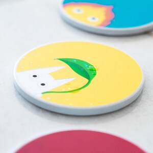 Puede incluir: Un conjunto de posavasos redondos con dise&ntilde;os coloridos y caprichosos. Un posavasos presenta un fondo amarillo con una criatura blanca y una hoja verde con gotas de agua. Otros posavasos tienen fondos azules y rojos con ilustraciones de dibujos animados.