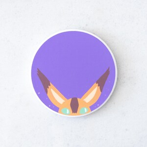 Puede incluir: Una pegatina redonda con un fondo morado y un dise&ntilde;o de zorro de dibujos animados. El zorro tiene orejas naranjas con puntas marrones, ojos verdes y una nariz marr&oacute;n. La pegatina tiene un borde blanco.