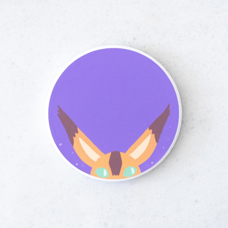 Puede incluir: Una pegatina redonda con un fondo morado y un dise&ntilde;o de zorro de dibujos animados. El zorro tiene orejas naranjas con puntas marrones, ojos verdes y una nariz marr&oacute;n. La pegatina tiene un borde blanco.