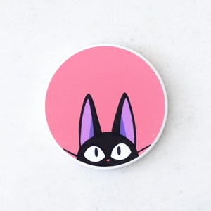 Puede incluir: Un soporte para tel&eacute;fono redondo y rosa con un gato negro con grandes ojos blancos y orejas forradas de p&uacute;rpura. La cabeza y las orejas del gato son el punto focal, asom&aacute;ndose por el borde inferior del c&iacute;rculo rosa.