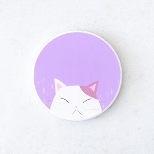 Puede incluir: Un posavasos redondo blanco con un dise&ntilde;o de gato de dibujos animados. El gato tiene pelaje blanco con una oreja rosa y una expresi&oacute;n de mal humor. El fondo es de color lila claro con peque&ntilde;as ilustraciones de peces.