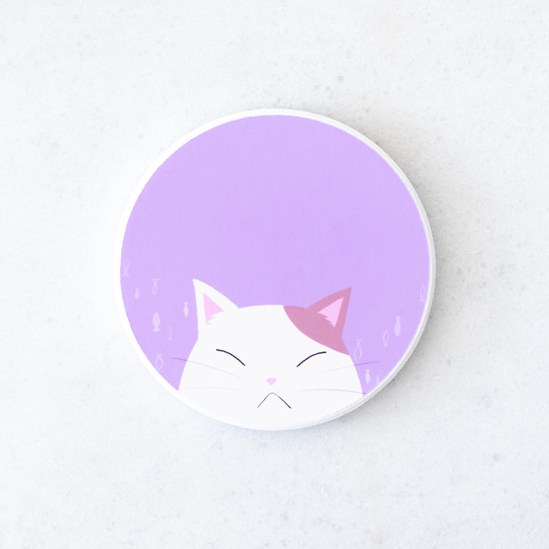 Puede incluir: Un posavasos redondo blanco con un dise&ntilde;o de gato de dibujos animados. El gato tiene pelaje blanco con una oreja rosa y una expresi&oacute;n de mal humor. El fondo es de color lila claro con peque&ntilde;as ilustraciones de peces.