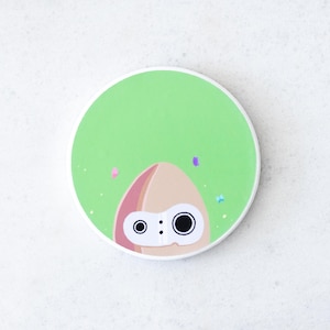 Puede incluir: Un posavasos redondo, verde y blanco, con un personaje de dibujos animados parecido a un calamar. El personaje tiene una cabeza marr&oacute;n claro con detalles rosas y dos grandes ojos negros. Peque&ntilde;as formas coloridas en forma de confeti est&aacute;n dispersas alrededor del personaje.