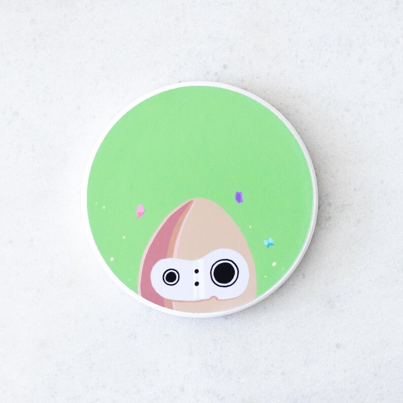Puede incluir: Un posavasos redondo, verde y blanco, con un personaje de dibujos animados parecido a un calamar. El personaje tiene una cabeza marr&oacute;n claro con detalles rosas y dos grandes ojos negros. Peque&ntilde;as formas coloridas en forma de confeti est&aacute;n dispersas alrededor del personaje.