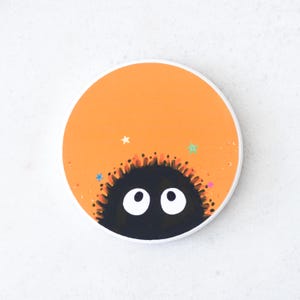 Puede incluir: Un posavasos redondo con fondo naranja y una criatura negra y peluda con ojos blancos que asoma por la parte inferior. El posavasos tiene un borde blanco y detalles de estrellas de colores.