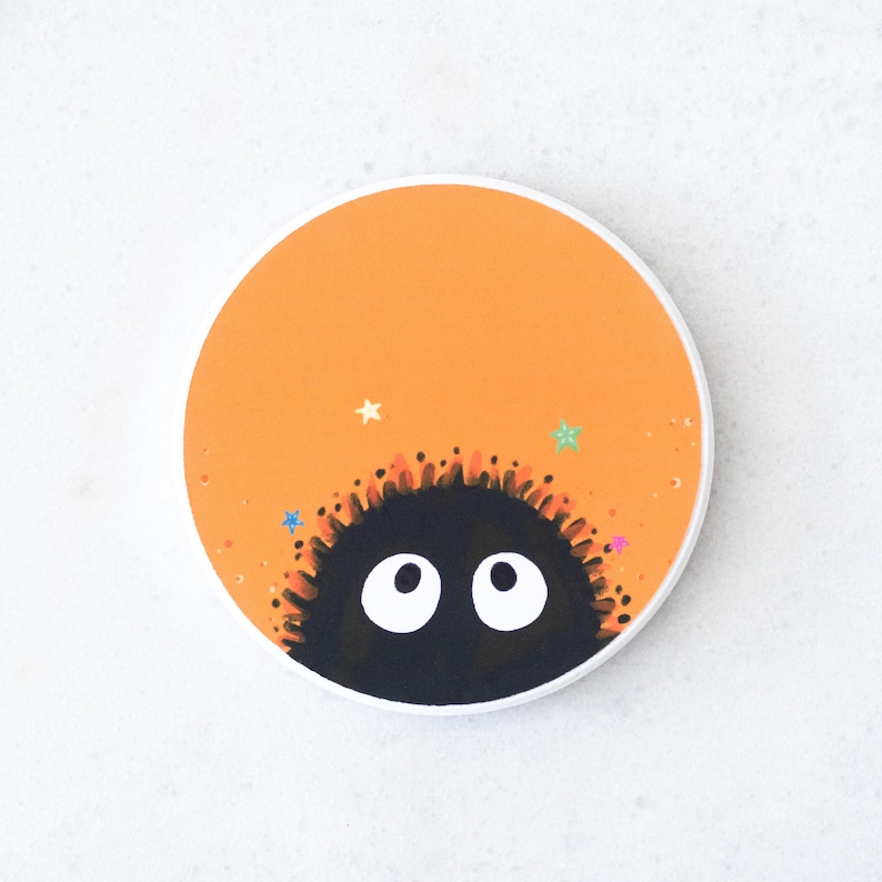 Puede incluir: Un posavasos redondo con fondo naranja y una criatura negra y peluda con ojos blancos que asoma por la parte inferior. El posavasos tiene un borde blanco y detalles de estrellas de colores.