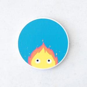 Puede incluir: Un posavasos redondo con un dise&ntilde;o de demonio de fuego de dibujos animados. El demonio tiene llamas amarillas con detalles en naranja y rojo, y dos ojos negros. El fondo es azul brillante, con un borde blanco.